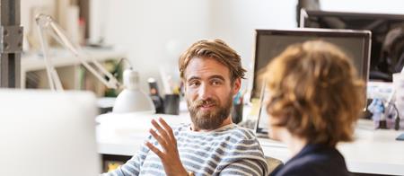 Two employees speaking while seated at computers